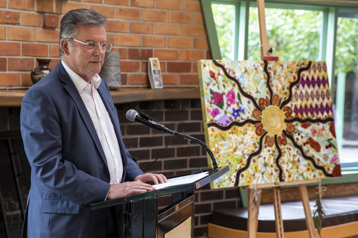 healthAbility Chair John Rasa stands and speaks in front of a seated audience. Next to him is an Aboriginal artwork on an easel