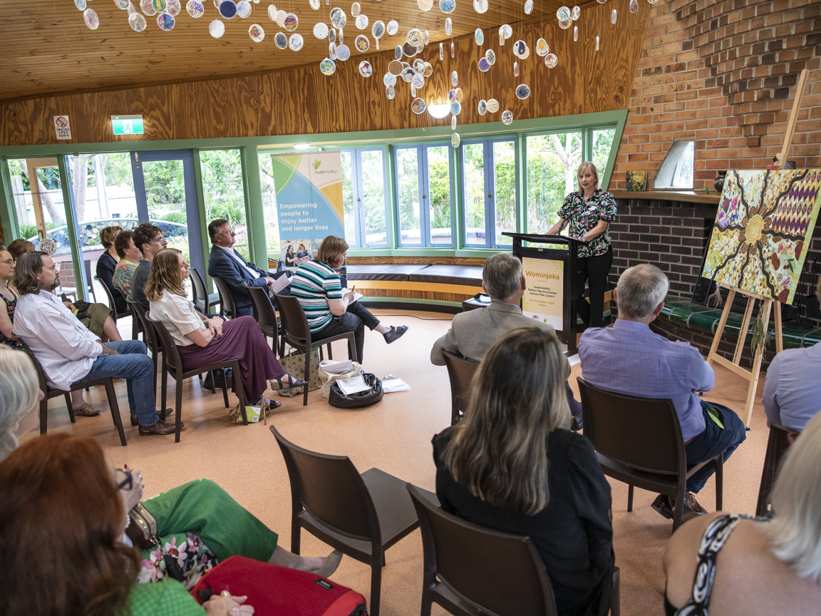 healthAbility CEO Agata Jarbin stands and speaks in front of a seated audience. Next to her is an Aboriginal artwork on an easel