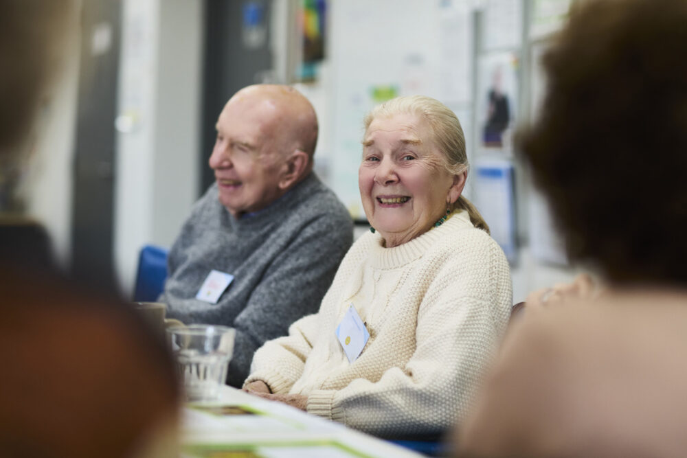 An older woman sitting next to an older man in a social activity group. She is smiling at the camera.